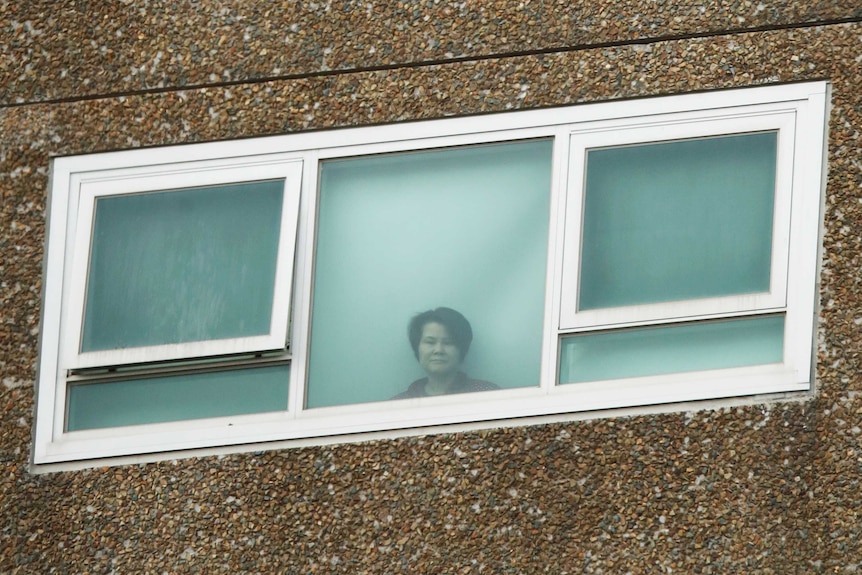 A woman looks out of a closed window in a tall brick building.