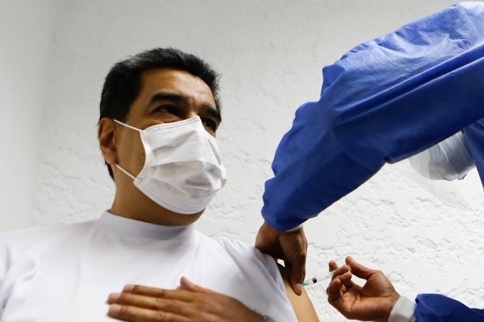 An elderly Hispanic man wearing a mask sits opposite a doctor in blue, giving him an injection.