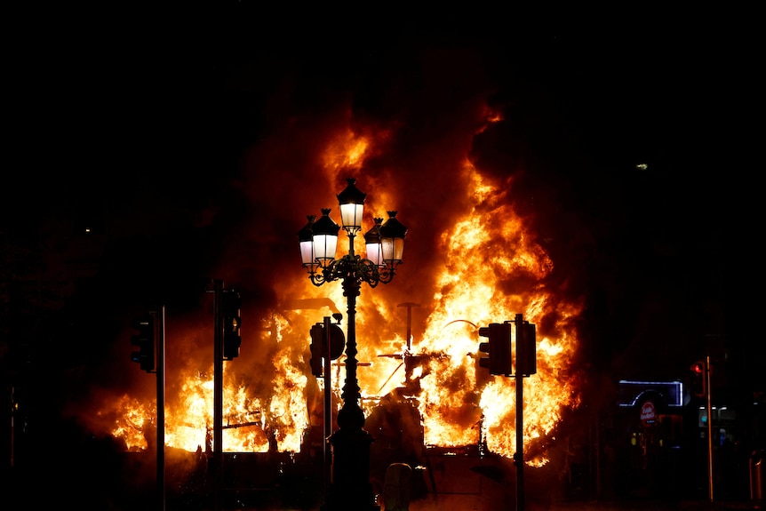 A bright fire burning during a dark night behind a lamp post and traffic lights