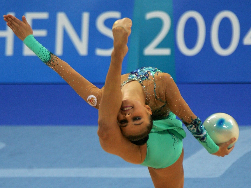 A rhythmic gymnast in turquoise leotard leans her head back towards a leg extended in the air, ball in one hand