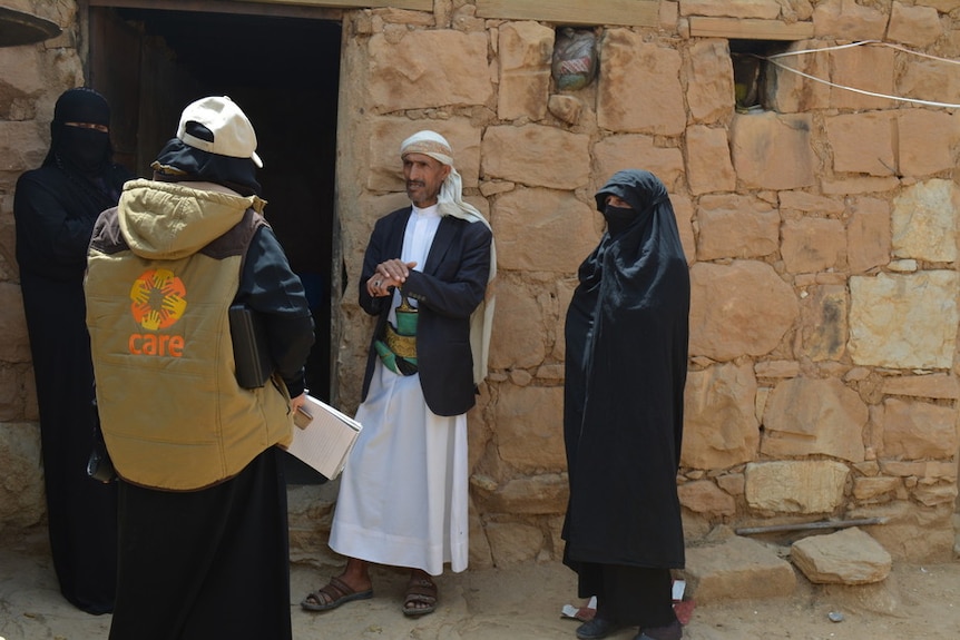 An aid worker speaks to an elderly man with a decorative knife in his belt who is flanked by two women.