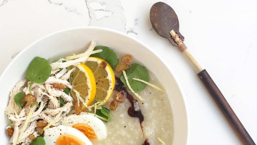 Arroz caldo soup with shredded chicken, lemon wedges, egg and other accompaniments in a white bowl alongside spoon.