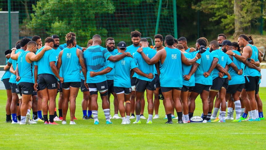 A group of Fijian Rugby players stand in a circle, arms interlocked and heads bowed.