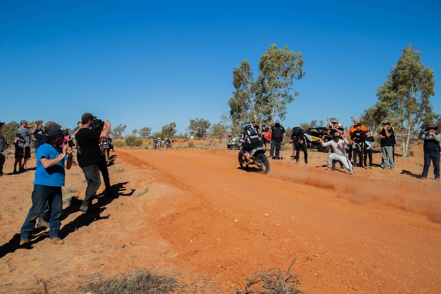 Spectators stand right on the edge of a desert race track as a motorcycle drives by.