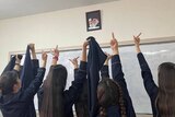 Five girls hold up their veils and make rude gestures to a hanging photo of the Iranian leader