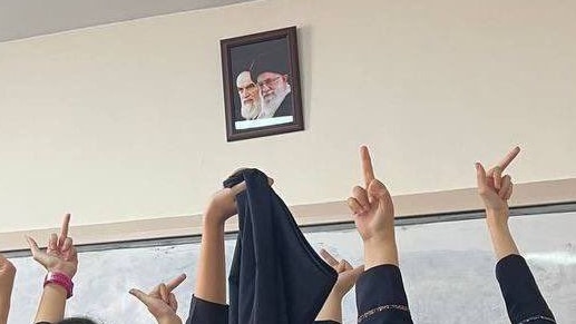 Five girls hold up their veils and make rude gestures to a hanging photo of the Iranian leader