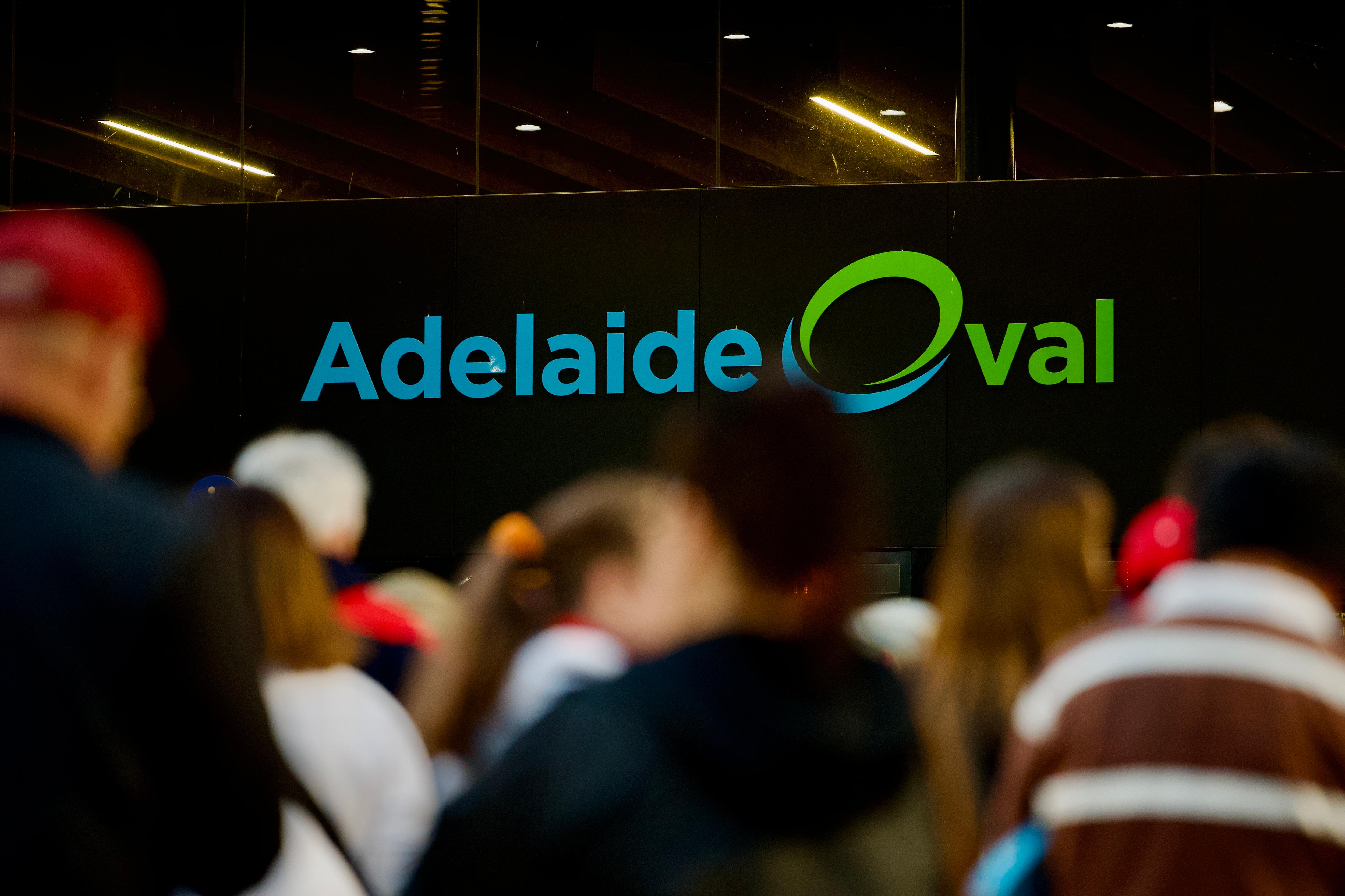 A parte de trás das cabeças caminhando em direção a uma placa iluminada que diz Adelaide Oval. 