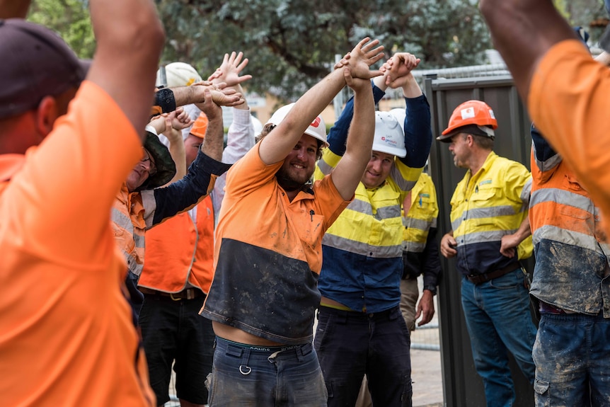 A group of men in orange high-vis stand and stretch, wearing hard hats.