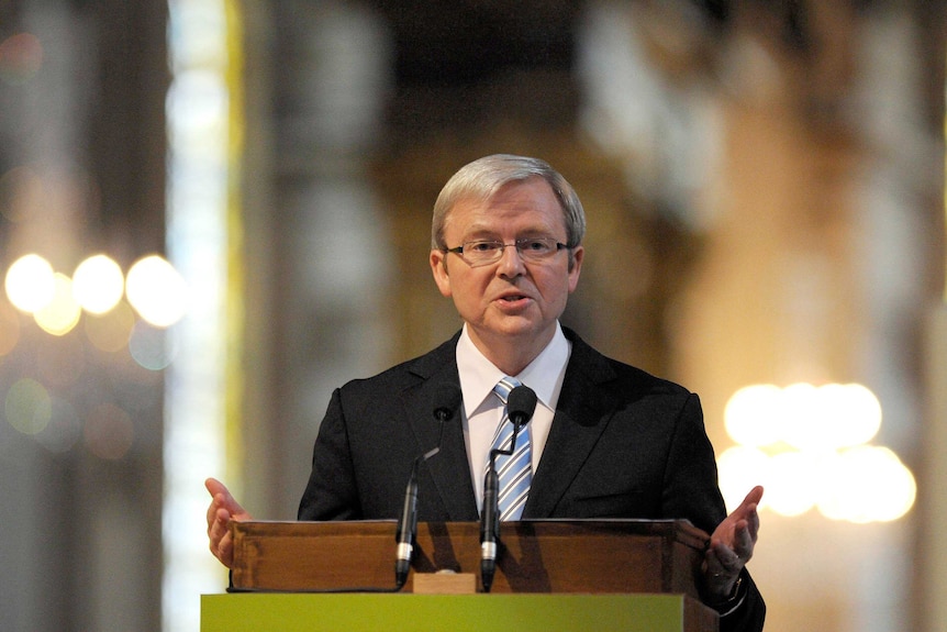 former PM kevin rudd stands in front of lectern gesturing with arms in front of blurred background featuring multiple candles