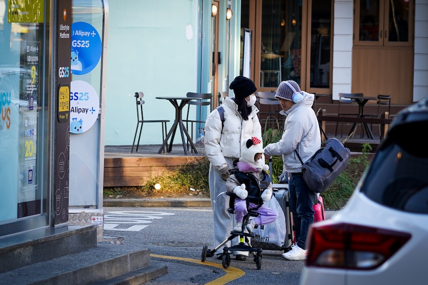 Two parents in winter coats stand on a street corner with their child and some luggage