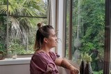A woman sits on her bed staring blankly out the window at green trees and ferns