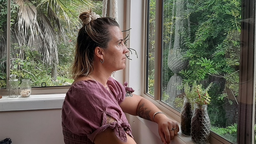 A woman sits on her bed staring blankly out the window at green trees and ferns