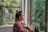 A woman sits on her bed staring blankly out the window at green trees and ferns