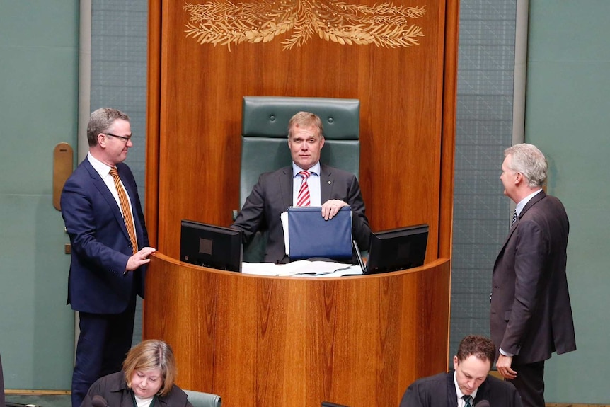 Tony Smith sits in the Speaker's chair, raising one eyebrow. Christopher Pyne and Tony Burke stand on each side of him