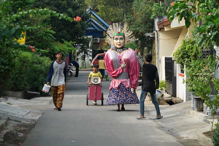 Perumnas Klender in East Jakarta. A person in an elaborately painted mask and costume is walking down a narrow alleyway.