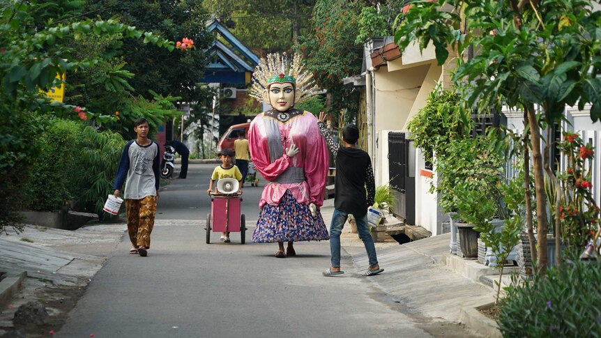 Perumnas Klender in East Jakarta. A person in an elaborately painted mask and costume is walking down a narrow alleyway.
