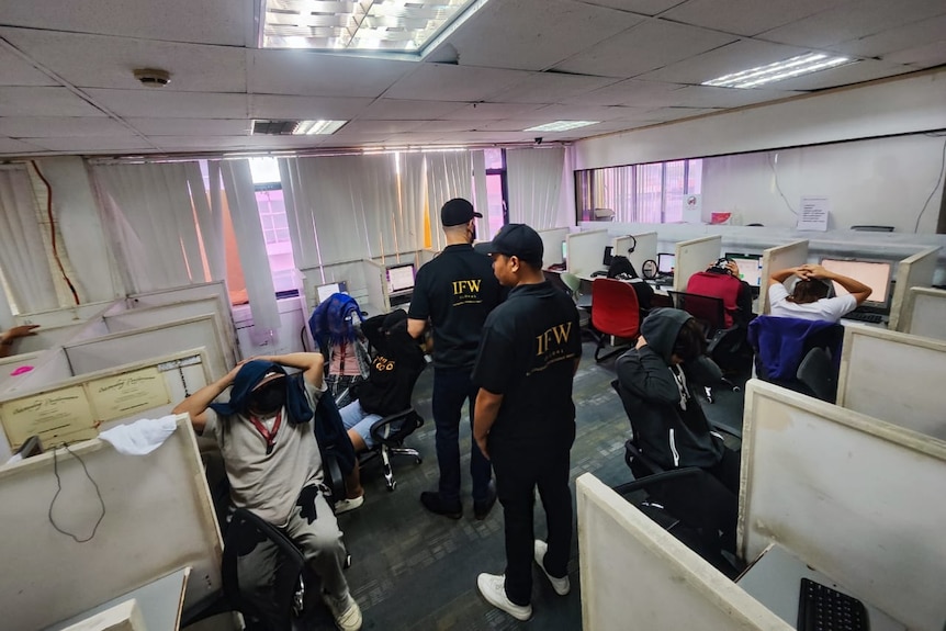 Investigators in an office with scam workers holding their hands on their heads.