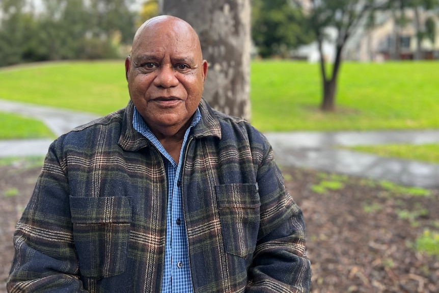 Bobby Nicholls smiles as he stands in a park, dressed in a warm checked jacket on a rainy day.