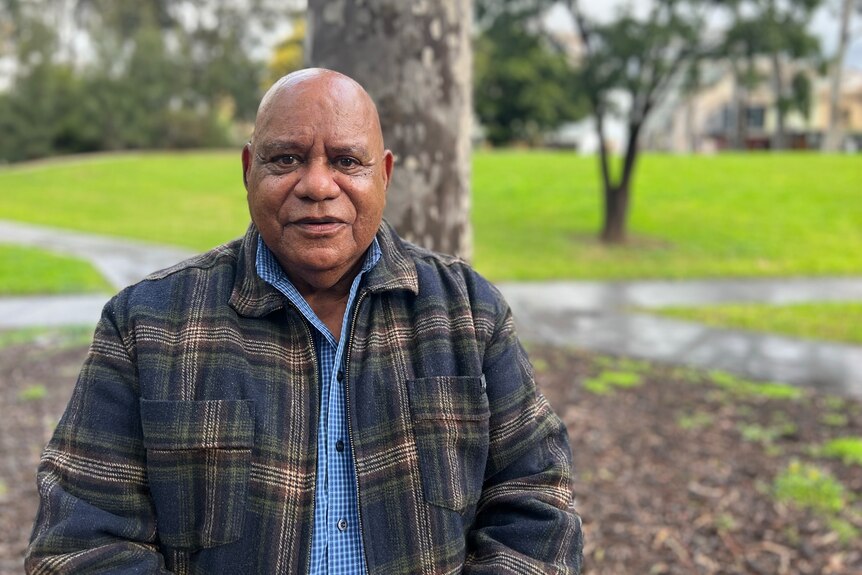 Bobby Nicholls smiles as he stands in a park, dressed in a warm checked jacket on a rainy day.