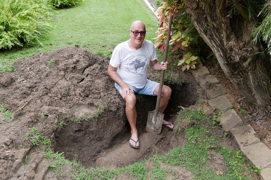 A bald man wearing sunglasses holding a shovel, sitting at the edge of a hole, part of a cannon visible in the dirt