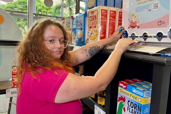 a woman in pink top reaches up to a tampon dispenser at a food bank