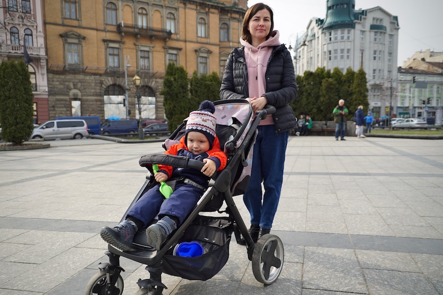 A woman wearing a pink jumper and puffer jacket leans on a pram carrying a boy wearing a beanie