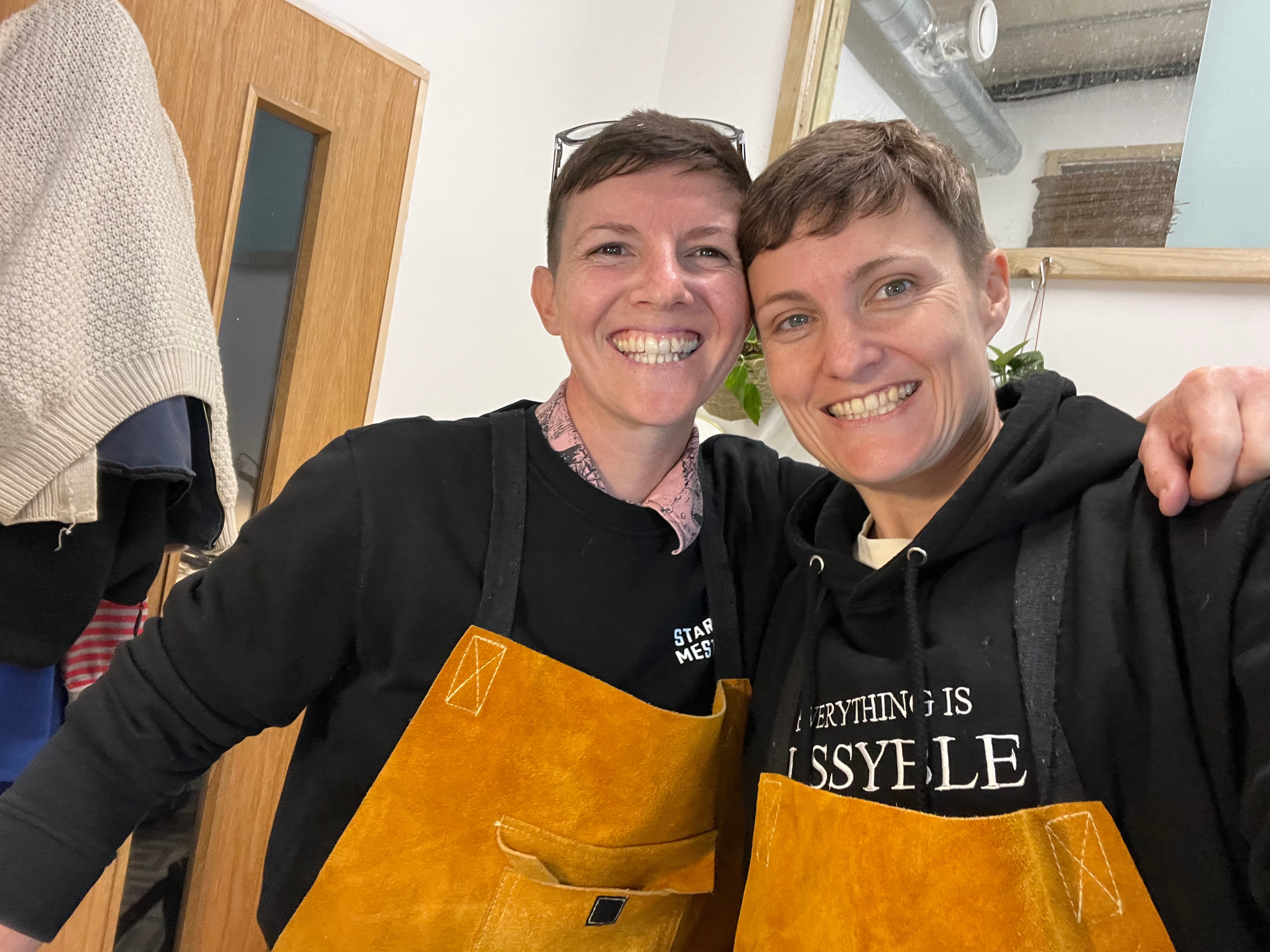 A female same-sex couple wearing black jumpers and aprons smile at the camera. The two women look very similar.