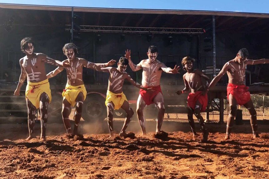 Six Indigenous men and boys perform on a red dirt dance ground.