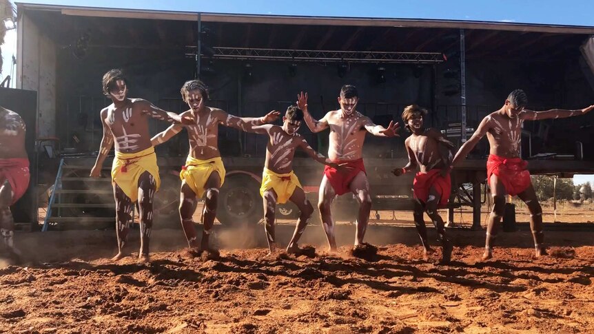 Six Indigenous men and boys perform on a red dirt dance ground.