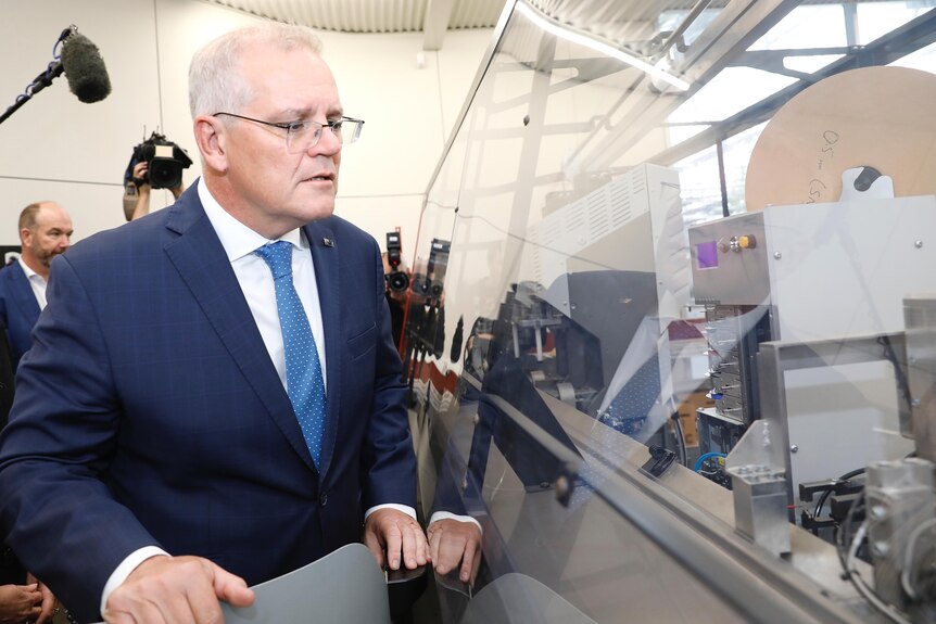 Scott Morrison looks at machinery through a glass panel with reporters behind him.