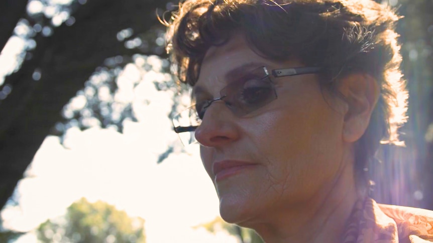 A middle-aged white woman with short curly hair and glasses. She is standing outside by some trees in the sun