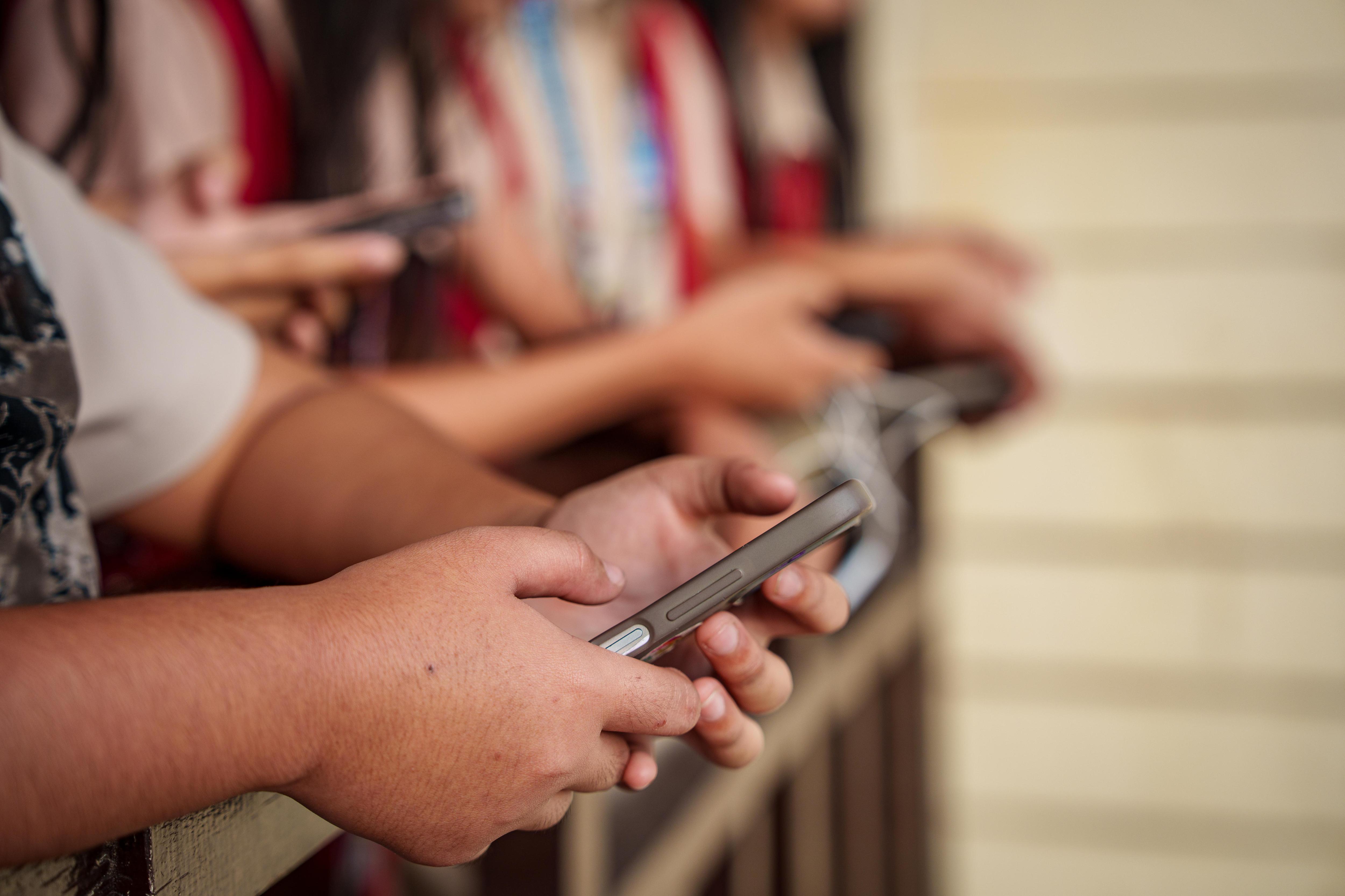 The hands of four school children holding their mobile phones while resting their forearms on a balcony railing.