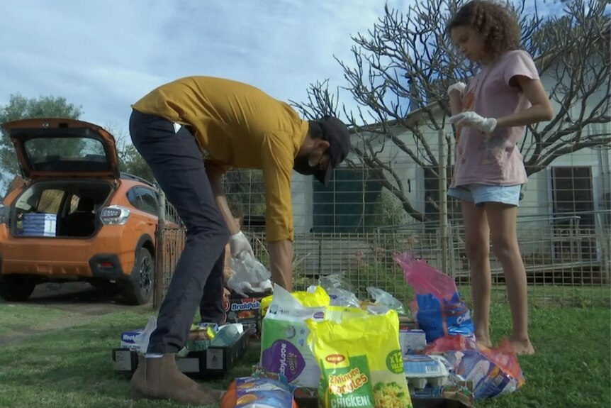 A man wearing a yellow shirt and a young girl in a pink top stand over groceries on the lawn outside of house.