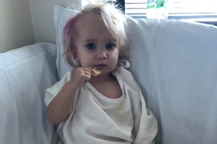 Cute blonde-haired baby sitting on hospital bed.