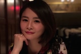 A woman sits in front of a table with her right hand holding her chin.