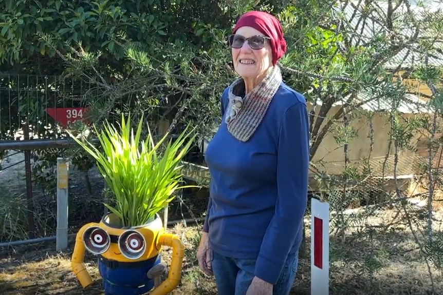 A woman with a red head covering and sunglasses stands next to her minion, it has a plant growing out of it like hair.