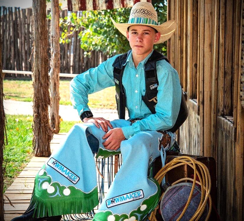 A young cowboy wearing a straw cowboy hat, blue shirt and jeans and brown vest.