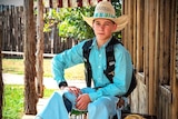A young cowboy wearing a straw cowboy hat, blue shirt and jeans and brown vest.