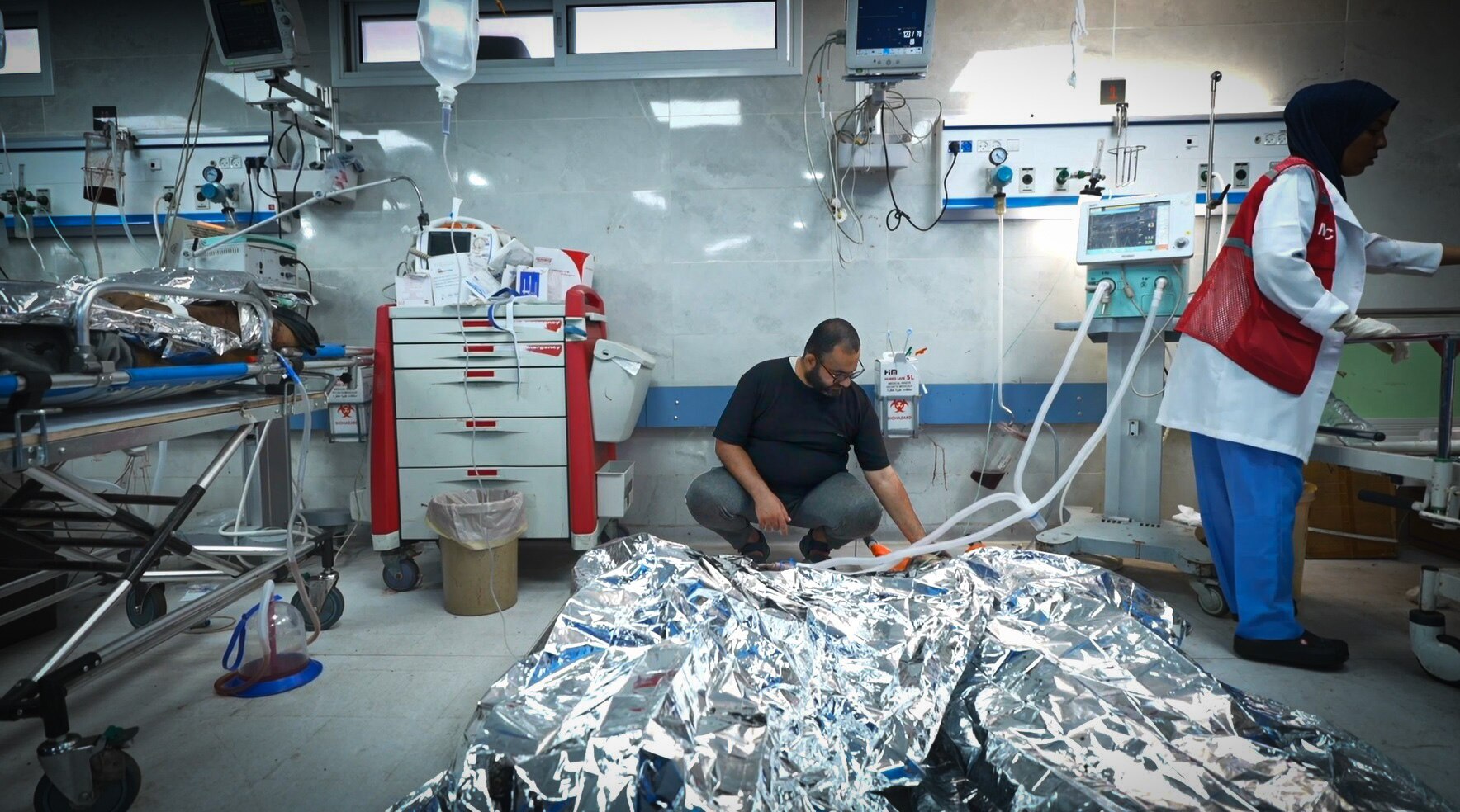 A man crouches on a hospital floor looking at people under silver blankets