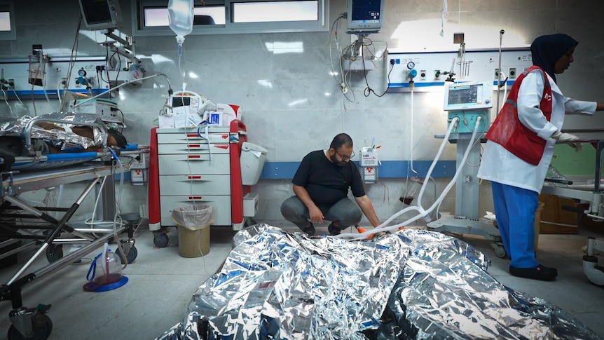 A man crouches on a hospital floor looking at people under silver blankets