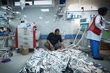 A man crouches on a hospital floor looking at people under silver blankets