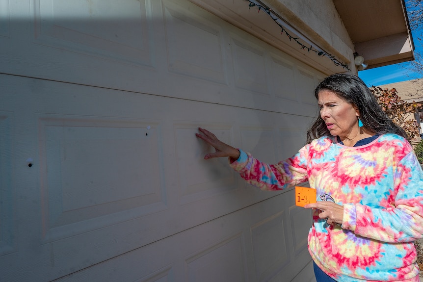 A woman in a brightly-coloured top surveys bullet holes in her garage door.