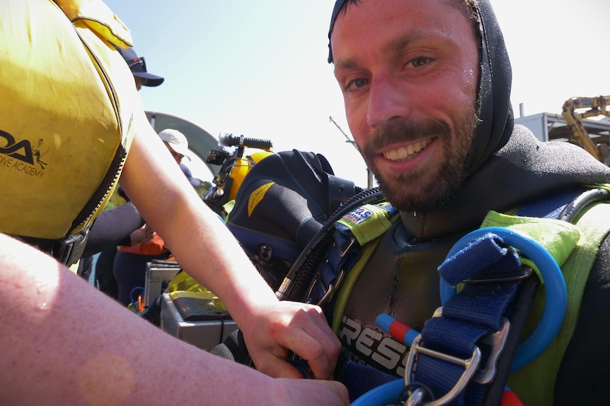 A man in a wetsuit and diving gear smiling to camera as someone helps remove his equipment.