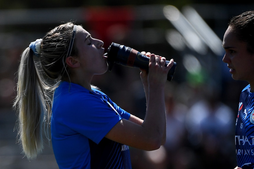 A soccer player wearing blue squirts water from a bottle into her mouth before a game