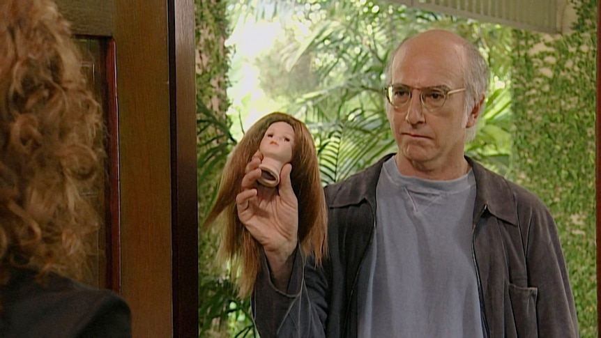 Larry stands in a doorway and holds the head of a brunette doll up with a serious expression on his face.