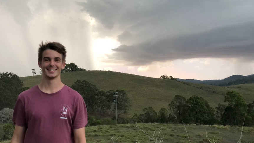 A young man stands, smiling for a photo, with  dramatic cloud formation behind him.