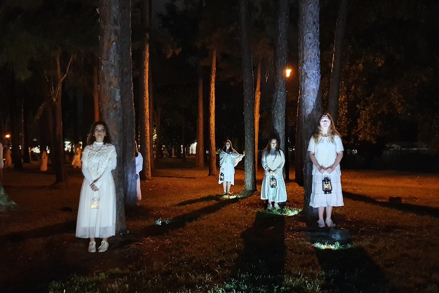 Girls in white stand in front of trees at night, hold lanterns, light projected on them, otherwise darkness.