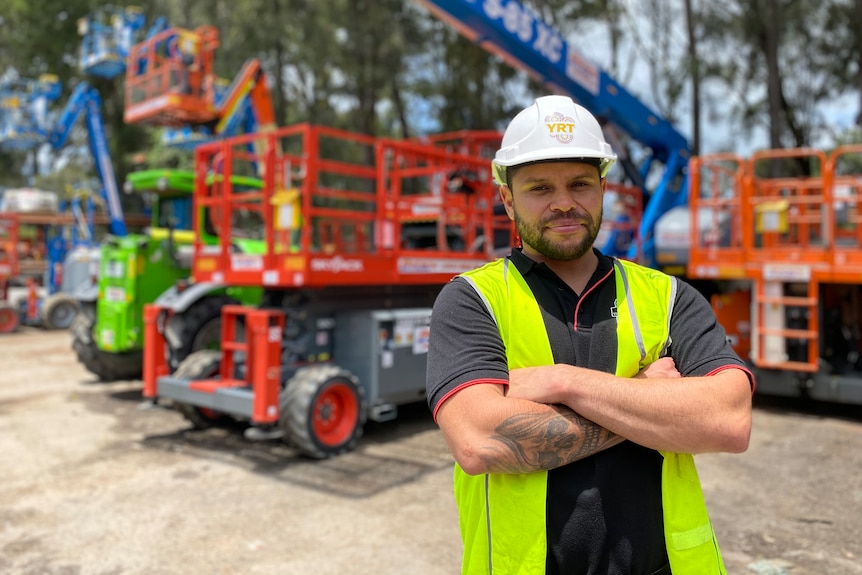 a man wearing a hard hat and high viz vest with arms folded standing in front of machinery