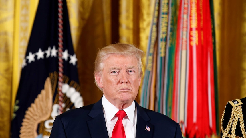 Donald Trump stands in front of a bright flags with a serious expression on his face.