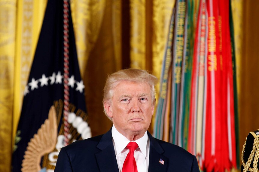 Donald Trump stands in front of a bright flags with a serious expression on his face.